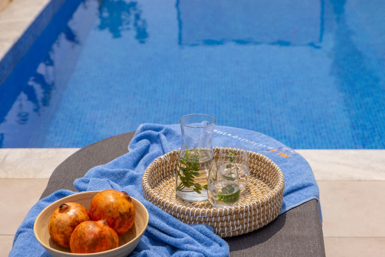 A woven tray of water and mint set beside a bowl of pomegranates at the edge of the pool.